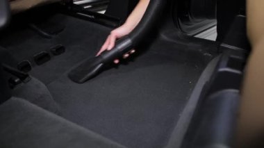 Arabanın iç tarafındaki halı üzerindeki çöpleri elektrikli süpürge kullanarak temizliyorum. Bir kadın eli hortumu kontrol eder. Siyah iç ve aydınlık koltuklar.