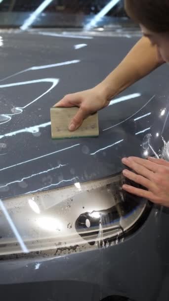 垂直录像 这名工人是一名妇女 她在一个灰色的新车车体上贴上防砂薄膜 并在详细的车辆维修车间放置废料 带特殊薄膜的车体保护概念 — 图库视频影像