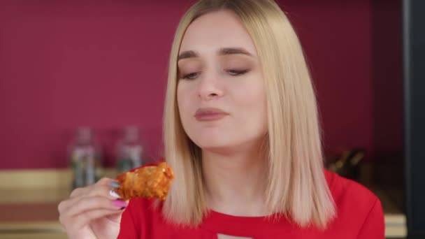 キッチンの赤い黒い部屋の背景にある若い金髪の女性は それを味わっている間に鶏の翼を食べます ファーストフードのコンセプト — ストック動画