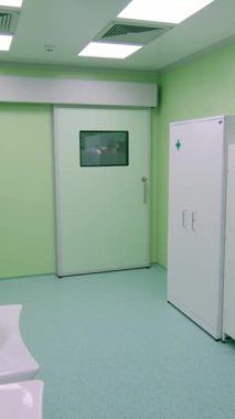 Dikey video. Ameliyathanenin önündeki boş koridorda bir dolap, lavabo ve havlular var. Ameliyat yapan doktorlar ve bir cerrah kapının penceresinden görülebilir. İlaç..