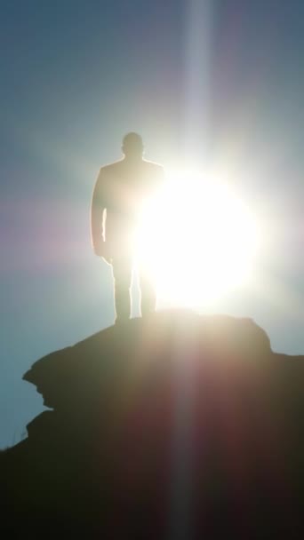 バーティカルビデオ 太陽に面した丘の上に立っている男の全長のシルエット 輝く円盤は 青空の背景に対して人体を取り囲んでいます 映画ドローンによる撮影 — ストック動画