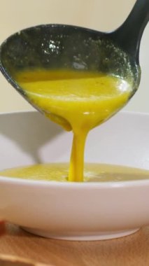 Dikey video. Kahverengi krema çorbası plastik siyah bir kepçeden beyaz bir tabağa dökülür. Hafif ahşap masa zemininde marul yaprakları, dereotu, ekmek kırıntıları..