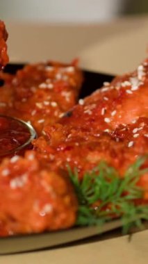 Dikey video. Kapatın. Parmaklar bir tavuk kanadını alır ve masanın üzerindeki ketçap kasesine batırır. Pişmiş fast food, dereotu ve susam tohumlu tabak..