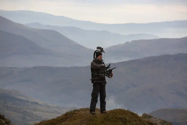Elinde siyah elbiseli bir kamerayla vahşi doğadaki bir dağın tepesinde arka planda dağlarla duran bir kameraman. Film endüstrisi ekipmanları ve işçiler.
