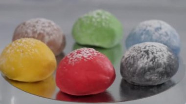 Altı kahverengi, yeşil, kırmızı, siyah, sarı ve siyah mochi kekleri ayna yüzeyinde yatarken dönüyorlar. Hafif bir arkaplanda pasta aromalarının sunumu.