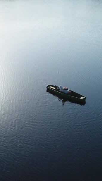 バーティカルビデオ 男は木製のボートに横たわりながらギターを演奏し 太陽の光が波に映し出される水の上の野生湖の真ん中にいる エアリアルビュー — ストック動画