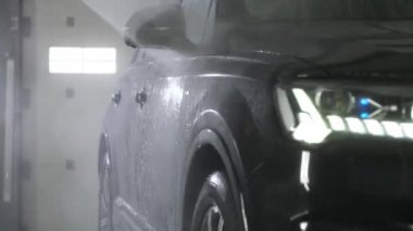 Bir oto yıkama işçisi, siyah bir otomobilin sağ tarafını yüksek basınçlı yıkama makinesiyle yıkıyor. Ön cam, çatı ve kapılar su püskürtüsünün altında. SUV bakım konsepti.