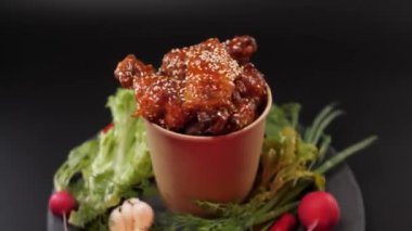 Baharatlı tavuk kanatları, üzerine susam tohumu serpiştirilmiş kağıt kovaya batırılmış sos ve taş bir tabakta sebzeler. Siyah arkaplan.