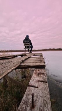 Dikey video. Koyu renk elbiseli bir adam, buzla kaplı bir göle bakan ahşap bir köprüdeki sandalyeye oturur. Akşam gökyüzünde hızlı hareket eden bulutlar. Yalnız devlet kavramı.