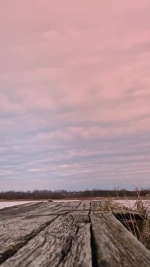 Dikey video. Donmuş bir gölün üzerinde karşı kıyıya bakan eski bir ahşap köprü. Akşam gökyüzünde bulutların hızlı hareketi.