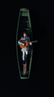 Dikey video. Gri montlu yetişkin bir adam ahşap yeşil otantik bir teknede yatıyor, gitar çalıyor ve şarkı söylüyor. Kayıkçının etrafı berrak ve karanlık sularla çevrili. Hava görünümü.