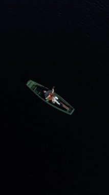 Dikey video. Bir adam, dalgalarda güneşin küçük yansımalarıyla vahşi bir gölün ortasında, ahşap bir teknede gitar çalar. Hava görünümü.