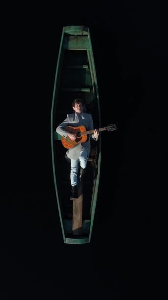 垂直录像 一个穿着灰色外套的成年男子躺在一艘木制绿色正宗的船上 弹吉他 船夫周围都是清澈的黑水 空中景观 — 图库视频影像