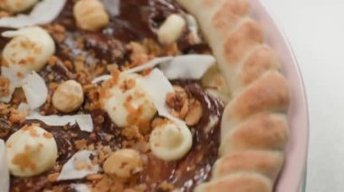 Kapatın. Beyaz arka planda pembe bir tabakta çikolatalı pizza maskarpon, fındık, hindistan cevizi talaşı ve krema karışımı içerir..