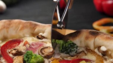 Yuvarlak pizza bıçağı taze bir pizzanın yüzeyinde yuvarlanır ve keser. Tekerlek vitrininde kırmızı biber, brokoli, peynir, jambon. Kapat..