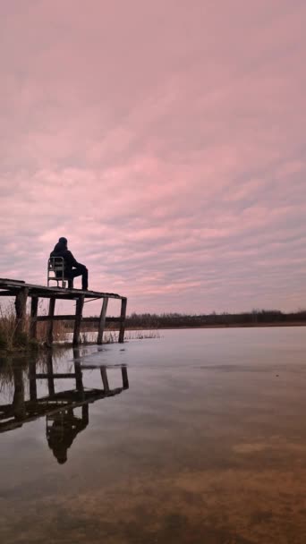 垂直录像 一个穿着保暖衣服的孤独男子坐在一座木桥上的椅子上 俯瞰着冰封的湖面 在夜空中快速移动的云彩 心理状态的概念 — 图库视频影像