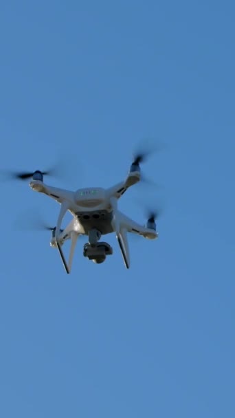 バーティカルビデオ 青い空の背景に飛行中の白いクワッドコプター フレーム内のドローンの外観とフレームからの出発 — ストック動画