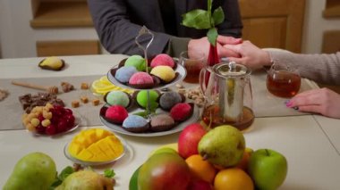 Bir erkeğin eli, bir kadının elini nazikçe mutfak masasında tutar. Bir tatlı mochi, mango, armut, elma, ahududu, kiraz, şam fıstığı, limon ve çay ile servis edilir..