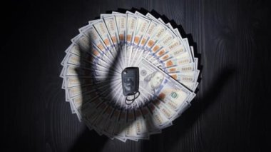 Tahta bir masa yüzeyinde daire şeklinde yığılmış yüz dolarlık banknotlar. Paranın ortasında, arabadan alınmış bir anahtarlık var. Ellerin gölgeleri tüm parayı ve anahtarı almaya çalışır..