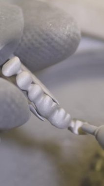 Dikey video. Diş teknisyenleri yapay seramik dişleri diş laboratuarında kum püskürtücü kullanarak cilalıyor. Profesyonel eldiven giyer, sahte dişleri elinde tutar ve kumla parlatır.