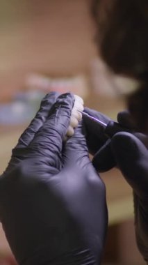 Dikey video. Diş teknisyeninin arka görüşü atölyesindeki seramik protezleri parlatıyor. Çerçevede ustanın siyah eldivenlerindeki eller ve uzman tarafından yaratılan dişler var..