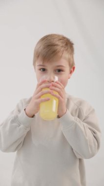 Dikey video. Beyaz, mavi gözlü sarışın çocuk bardaktan limonata içer ve kendini yalamaya başlar. Beyaz arka plan. Sağlıklı doğal gıda kavramı.