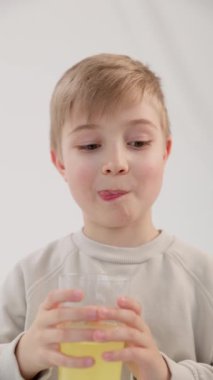 Dikey video. Mavi gözlü, limonata kadehiyle gülümseyen ve dudaklarını lezzetli içecekten yalayan sarışın beyaz bir çocuk. Beyaz arka plan. Sağlıklı doğal gıda kavramı.