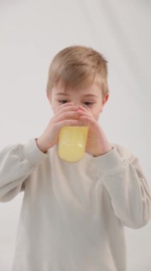 Dikey video. Beyaz, mavi gözlü sarışın çocuk bardaktan limonata içer ve kendini yalamaya başlar. Beyaz arka plan. Sağlıklı gıda kavramı.