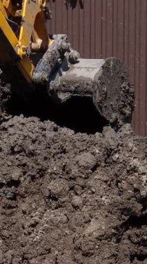 Dikey video. Bir traktör kovası bir yaz günü özel bir evin kahverengi çitlerinin yanında çukur kazar..