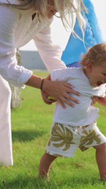 Dikey video. Küçük çocuk annesinin kollarından kurtulur ve çimlerin üzerinde yürür. Evin yanında yaz yürüyüşü. Çocuklukta bağımsızlık ve azim kavramı.