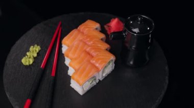 Somon, wasabi ve zencefilli Philadelphia suşisi seti taştan bir tabakta siyah arka planda döner. Tabağın yanında kırmızı yemek çubukları ve bir kavanoz soya sosu var..