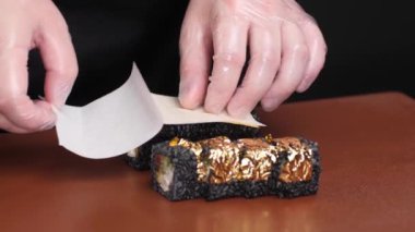 Şeflerin elleri şeffaf eldivenlerle siyah suşi takımına altın takıyor. Tabağa mürekkep balığı mürekkebi, peynir, somon ve mango turşulu siyah pirinç de dahil..