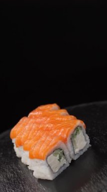 Dikey video. Somon, wasabi ve zencefilli Philadelphia suşisi seti taştan bir tabakta siyah arka planda döner. Boşluğu kopyala.