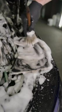 Dikey video. Kapatın. Siyah eldivenli bir el, fırçayla bir arabanın kaputunun altındaki motor elementlerini yıkar ve temizler. Sabun köpüğü ve deterjan. Araba temizleme kavramı.