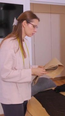 Dikey video. Bir kadın elbise dikmek için kumaş üzerinde bir desen inceler. Boynunda bir mezura, beyaz tuğla bir duvar ve masanın üzerinde dikiş makinesi olan bir odada..