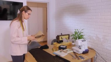 Bir kadın elbise dikmek için kumaş üzerinde bir desen inceler. Boynunda bant olan pembe bir kazak beyaz tuğladan bir duvar ve masanın üzerinde dikiş makinesi olan bir odada..