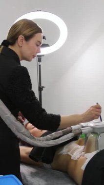 Dikey video. Modern bir kozmetoloji kliniğinde kriyolipoliz yağ tedavisi prosedürü. Siyah elbiseli bir kadın uzman, zayıflama aleti olan bir hastaya hizmet eder..