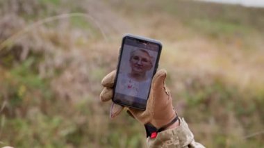 Sağ el yakın plan askeri taktik eldivenli akıllı telefon ve gözlüklü yaşlı bir kadının fotoğrafı. Sevdiklerinin anıları. Vücut parçası Ukrayna ordusu.