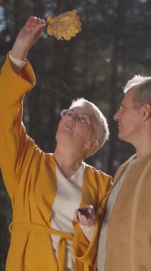 Dikey video. Huzurlu bir ormanda, yaşlı bir çift, ağaçlarla çevrili altın saat boyunca aşklarını kutlarken kalpten bir öpücük paylaşıyor. Evlilik ilişkileri