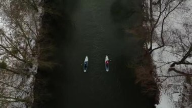 Hava görüntüsü. İki kanocu, karla kaplı kıyılar ve yapraksız ağaçlarla çevrili sakin bir nehirde kürek çekerek huzurlu bir kış ortamı yaratıyor. Aktif spor eğlencesi.