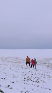 Dikey video. Üç kişilik bir aile, beyaz tarlalar ve bulutlu bir gökyüzü ile çevrili engin karlı bir arazide birlikte yürür. Baba yol gösteriyor, rahat bir ortam yaratıyor..