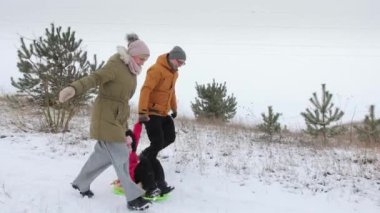 Genç karı ve koca, kızlarıyla el ele tutuşuyorlar. Plastik bir kızağa biniyor ve ailesi kızı karlı bir tepeden aşağıya taşımaya başlıyor. Kış eğlencesi başladı.