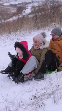 Dikey video. Bir ailenin plastik kızaktan aşağı inerken eğlendiği bir kar kaydırağı. Anne, kız ve baba oturur ve daha hızlı gitmek için kendilerini zorlarlar. Doğal tepelerde kış oyunları..