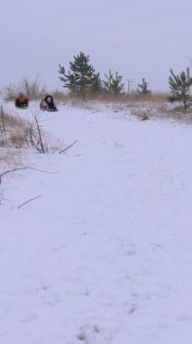 Dikey video. Anne ve kızı plastik kızaklarla kar kaydırağında kayarken eğleniyorlar. Babam çok geride. Aile kışı aktif eğlence.