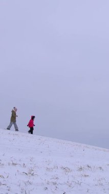 Dikey video. Genç bir aile, karlı bir yamaçta eğleniyor, oynuyor ve gülüyor, kış havasında bir tarlada koşuyorlar. Sonsuz beyaz manzara. Neşeli çocukluk ve aile değerleri.