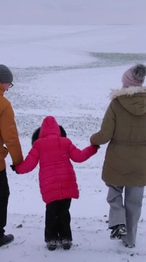 Dikey video. Üç kişilik bir aile, beyaz tarlalar ve bulutlu bir gökyüzü ile çevrili engin karlı bir arazide birlikte yürür. Kırmızılı bir çocuk öne atlıyor, neşeli anlar yaratıyor. Kış doğası