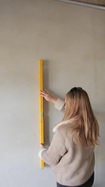 Dikey video. Genç bir kadın, yeni apartman inşaatı sırasında duvar yüzeyini sarı kabarcık seviyesiyle kontrol ediyor..
