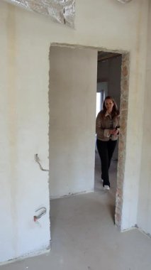 Dikey video. Bir kadın yeni bir evin koridorunda yürüyor ve tadilat yapılmış bir odaya giriyor. İnşaatın bir sonraki aşamasına hazırlanmak için alçıdaki gri tuğla duvarlara dokunuyor..