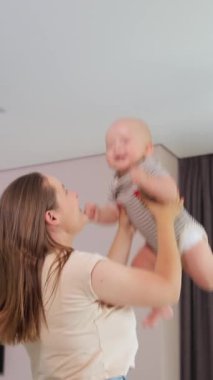 Dikey video. Uzun saçlı genç bir kadın, mutlu bir şekilde üst katındaki parlak bir odada, sade bir atmosferde bir bebeği fırlatıyor. Sevgi dolu bir anneyle neşeli bir çocukluk. Aile rahatlığı.