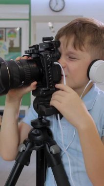 Dikey video. Açık mavi gömlekli genç bir çocuk parlak ışıklı bir sınıfta fotoğraf ve video çalışıyor, kulaklık takıyor ve kameranın merceğine odaklanmış ve heyecanlı bir şekilde bakıyor..